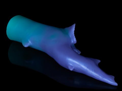Fantasy shark-inspired silicone stroker in a gradient of teal to purple, featuring subtle ridges, twin pelvic fins framing the entrance, and a long, pointed tail, displayed on a black background.