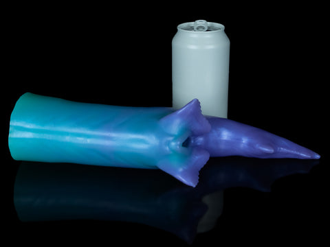 Fantasy shark-inspired silicone stroker in a gradient of teal to purple, featuring subtle ridges, twin pelvic fins framing the entrance, and a long, pointed tail, displayed on a black background. Next to the stroker is a grey soda can for scale.