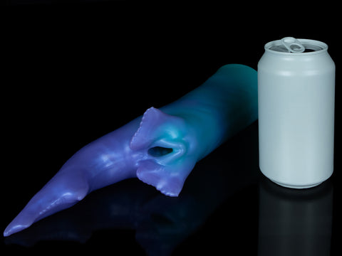 Fantasy shark-inspired silicone stroker in a gradient of teal to purple, featuring subtle ridges, twin pelvic fins framing the entrance, and a long, pointed tail, displayed on a black background. Next to the stroker is a grey soda can for scale.